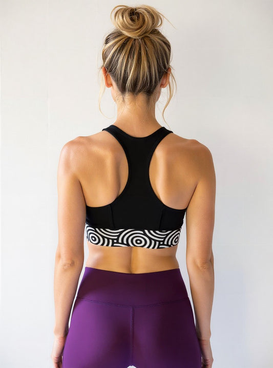 Athletic woman with blonde hair in a messy bun, shown from the back. She is wearing a black longline sports bra with a bold black and white circular "Lunar Loops" print band and high-waisted deep purple leggings, standing against a plain light background.