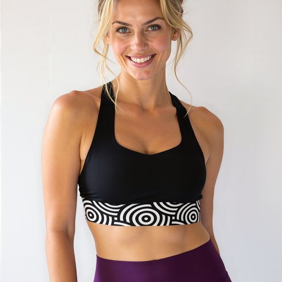 Smiling athletic woman with blonde hair in a messy bun, wearing a black longline sports bra with a bold black and white circular "Lunar Loops" print band, paired with high-waisted deep purple leggings, standing against a plain light background.