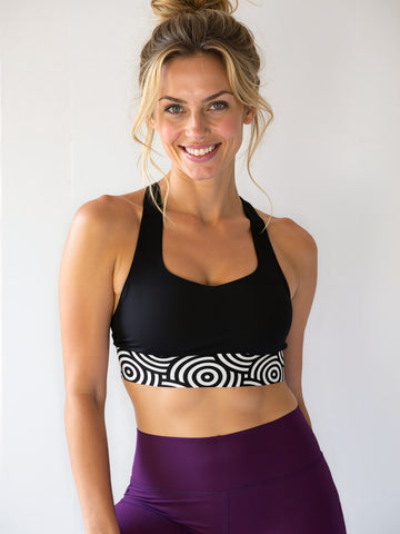 Smiling athletic woman with blonde hair in a messy bun, wearing a black longline sports bra with a bold black and white circular "Lunar Loops" print band, paired with high-waisted deep purple leggings, standing against a plain light background.