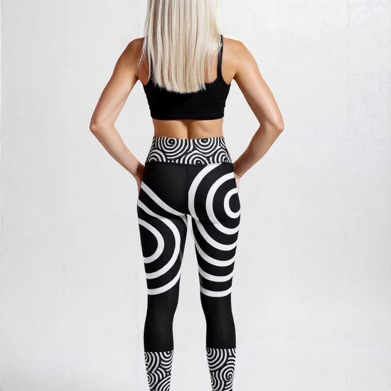 Back view of model wearing black high‑waisted Lunar Loops yoga leggings with bold white concentric circle and spiral pattern, paired with black sports bra and white sneakers, against a plain light background.