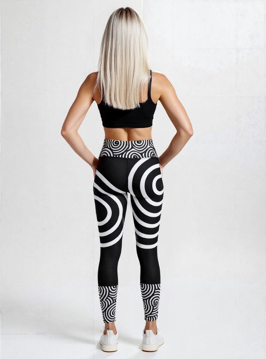 Back view of model wearing black high‑waisted Lunar Loops yoga leggings with bold white concentric circle and spiral pattern, paired with black sports bra and white sneakers, against a plain light background.