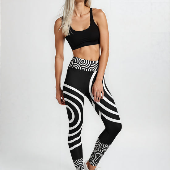 Woman wearing Lunar Loops Yoga leggings with a bold black and white concentric circle and swirl pattern, paired with a black sports bra and white sneakers, standing against a plain light background.