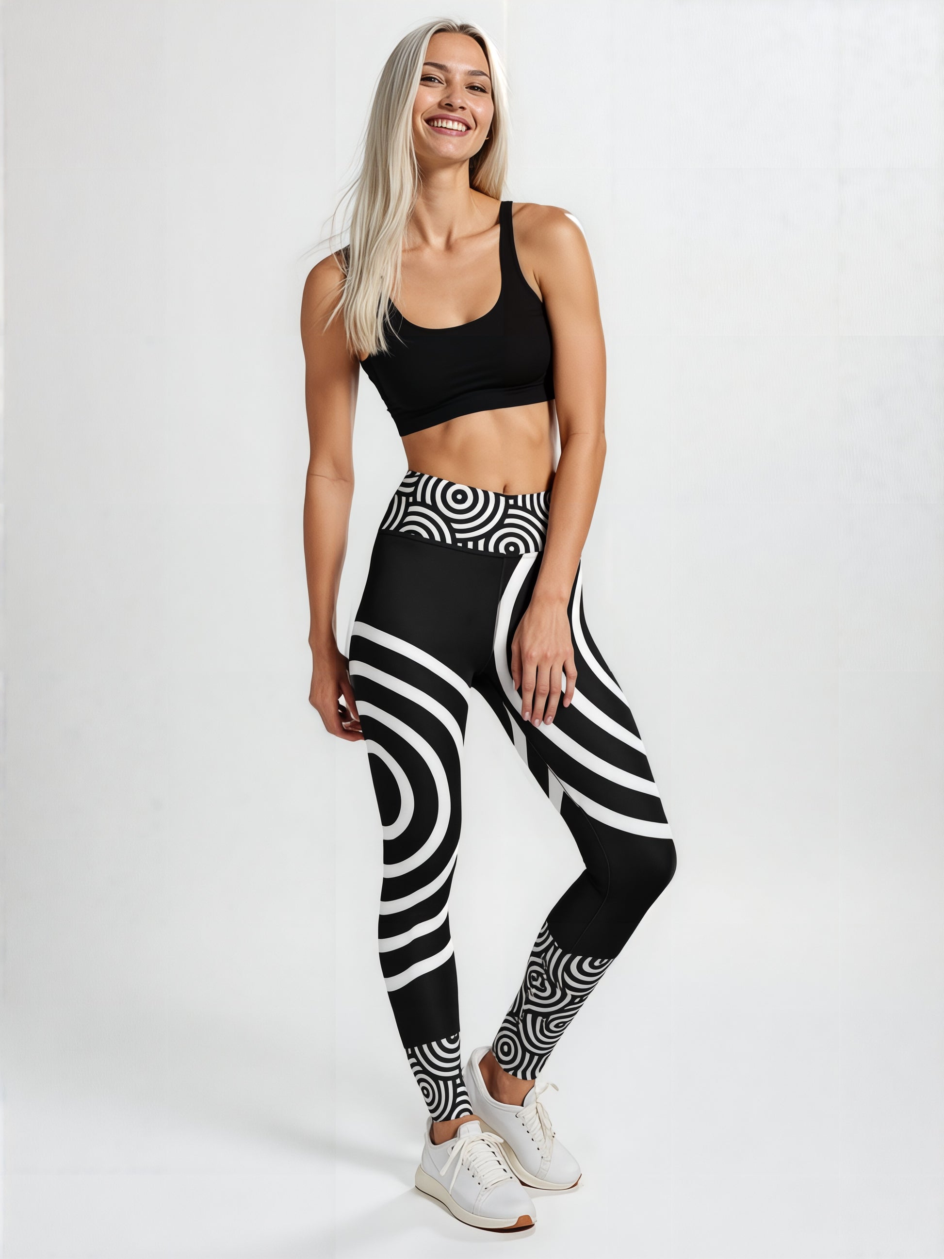Woman wearing Lunar Loops Yoga leggings with a bold black and white concentric circle and swirl pattern, paired with a black sports bra and white sneakers, standing against a plain light background.