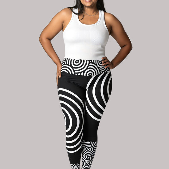 Woman wearing a white tank top and black and white patterned Lunar Loop Yoga leggings , by Pixie and Sherbet, on a plain background