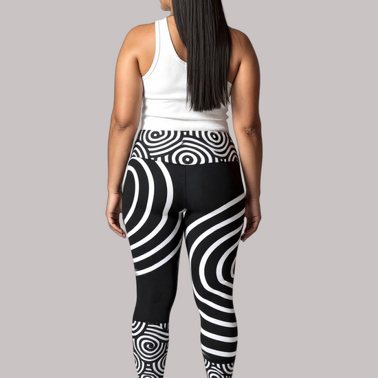 Back view of a model wearing Pixie & Sherbet’s Lunar Loops leggings with bold black and white concentric circle patterns, paired with a white sleeveless top and white sneakers, standing against a light background in a relaxed, upright pose.