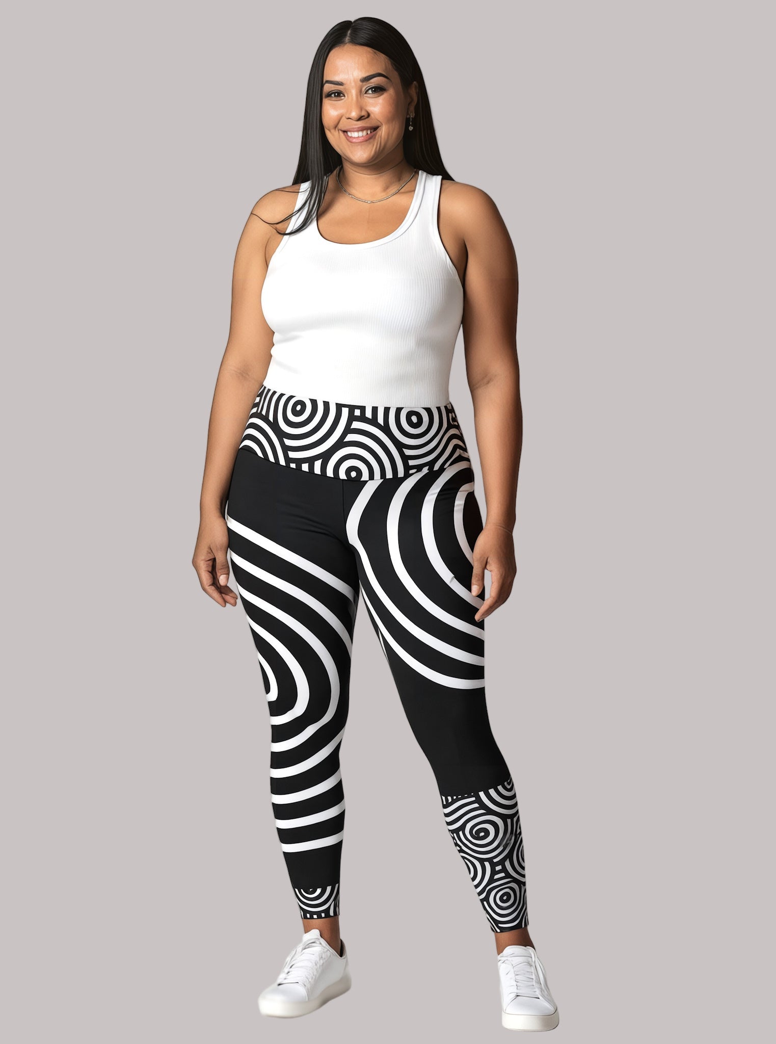 Model smiling in Pixie & Sherbet’s Lunar Loops leggings featuring bold black and white concentric circle patterns, paired with a white sleeveless top and white sneakers, standing confidently against a light background.