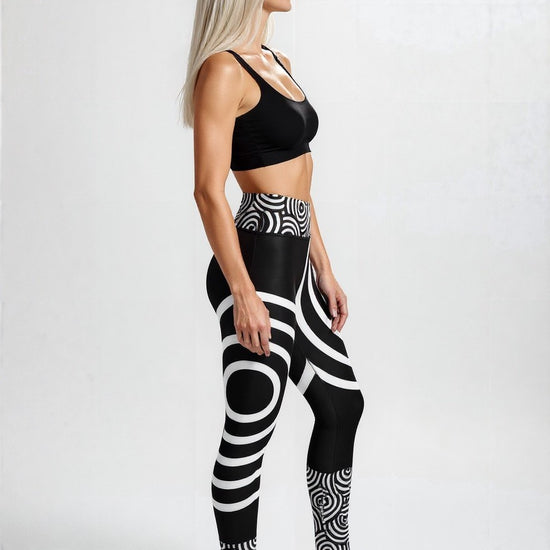 Side view of a woman wearing Lunar Loops Yoga leggings featuring a striking black and white geometric circle print, paired with a black sports bra and white sneakers, standing against a plain light background.