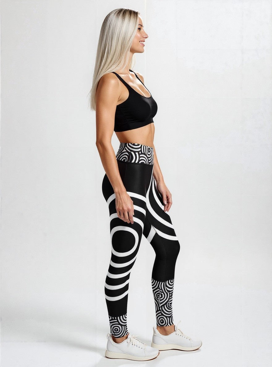 Side view of a woman wearing Lunar Loops Yoga leggings featuring a striking black and white geometric circle print, paired with a black sports bra and white sneakers, standing against a plain light background.