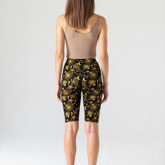 Back view of a woman wearing Midnight Halo bikers featuring a gold celestial pattern with stars, moons, and suns on a black background, paired with a beige tank top and black sneakers, standing against a plain background.