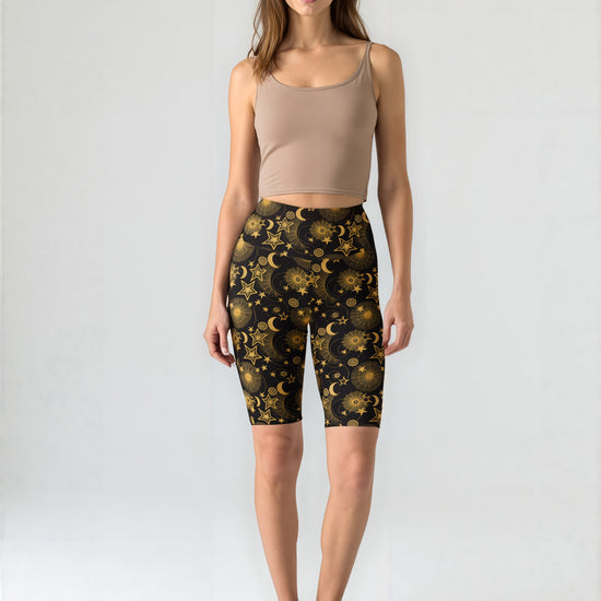 Woman standing and wearing Midnight Halo biker shorts with a black base and gold celestial star and moon print, paired with a beige crop tank top and black sneakers, against a plain background.