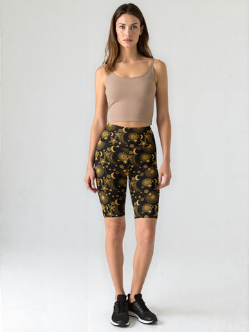 Woman standing and wearing Midnight Halo biker shorts with a black base and gold celestial star and moon print, paired with a beige crop tank top and black sneakers, against a plain background.