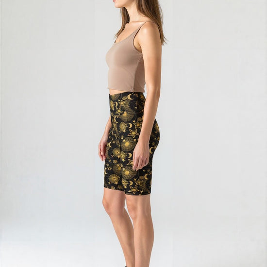 Side view of a woman wearing Midnight Halo biker shorts with a black base and gold celestial pattern of stars, moons, and suns, paired with a beige crop top and black sneakers, standing against a plain background.
