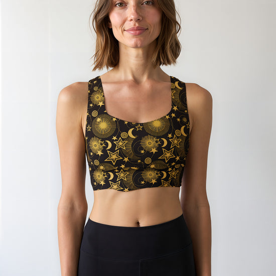 Woman wearing a Midnight Halo longline sports bra featuring a black base with intricate gold celestial designs, including stars, moons, and sunbursts, paired with matching high-waisted shorts, standing against a plain background.