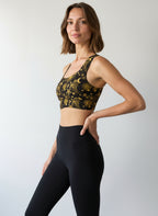 Woman wearing a Midnight Halo longline sports bra featuring a black base with intricate gold celestial designs, including stars, moons, and sunbursts, paired with matching high-waisted shorts, standing against a plain background.