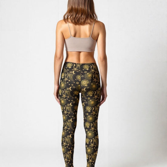Woman wearing Midnight Halo yoga leggings with a celestial pattern featuring gold stars, suns, and moons on a black background, paired with a beige sports bra, standing barefoot against a plain white backdrop.