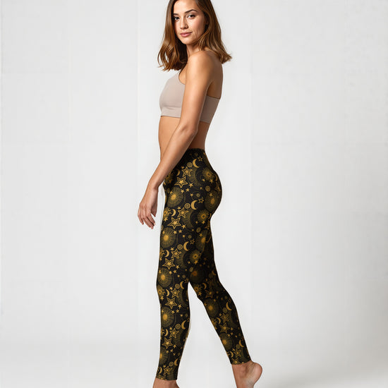 Side view of a woman wearing Midnight Halo yoga leggings featuring a celestial gold star, moon, and sun pattern on a black background, paired with a beige sports bra, standing barefoot against a plain backdrop.