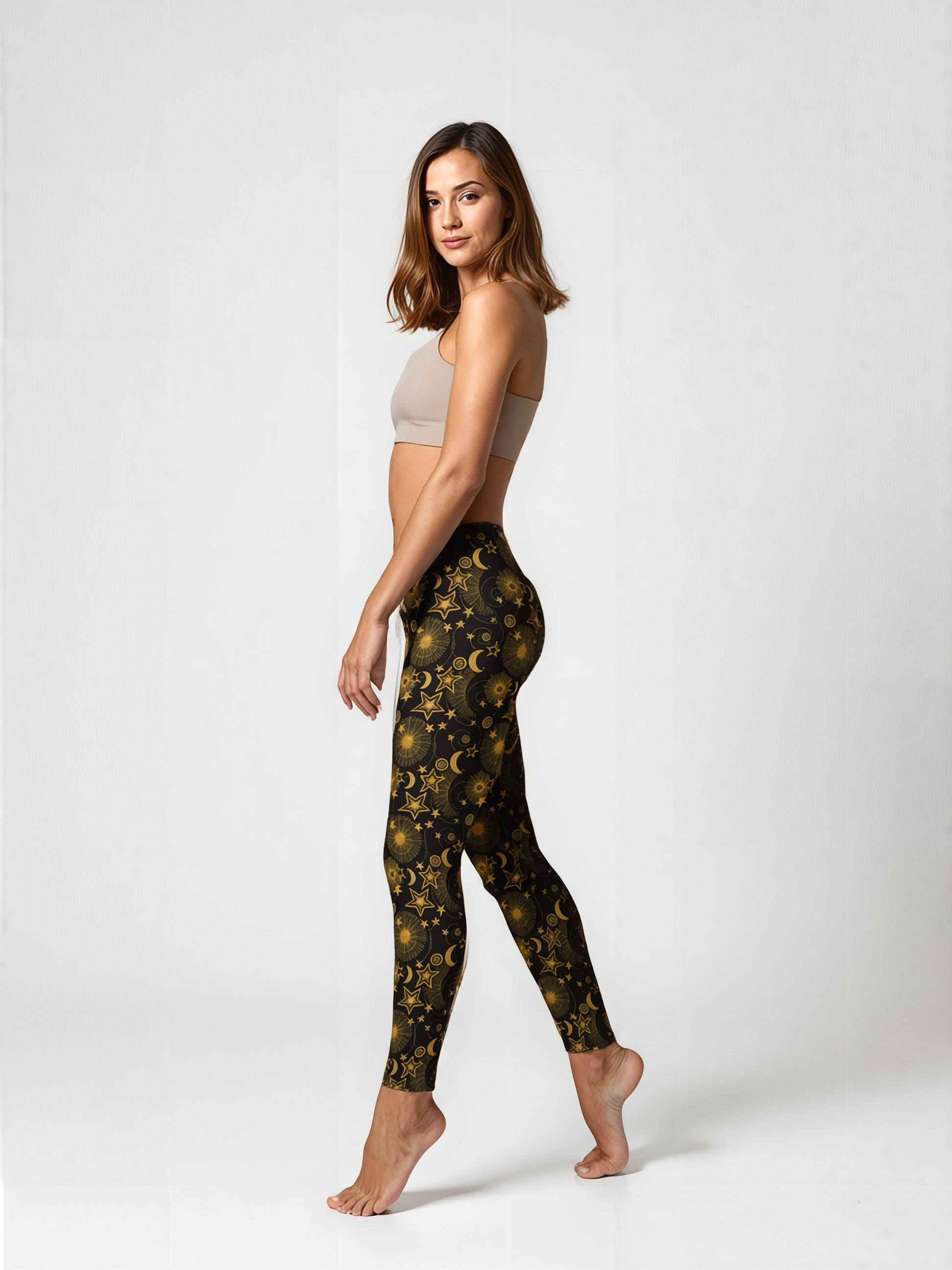 Side view of a woman wearing Midnight Halo yoga leggings featuring a celestial gold star, moon, and sun pattern on a black background, paired with a beige sports bra, standing barefoot against a plain backdrop.
