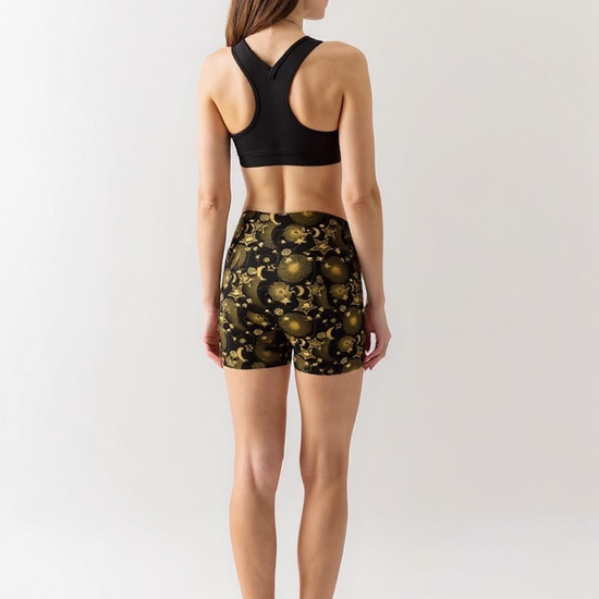 Woman wearing Midnight Halo yoga shorts with a black and gold celestial pattern of stars, suns, and moons, paired with a black sports bra, standing barefoot and turned slightly to the side against a plain light background.