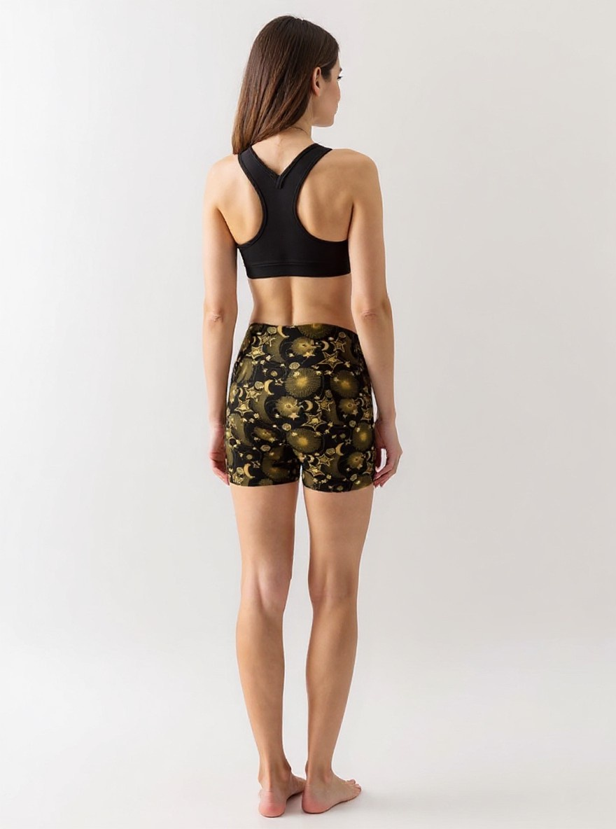 Woman wearing Midnight Halo yoga shorts with a black and gold celestial pattern of stars, suns, and moons, paired with a black sports bra, standing barefoot and turned slightly to the side against a plain light background.