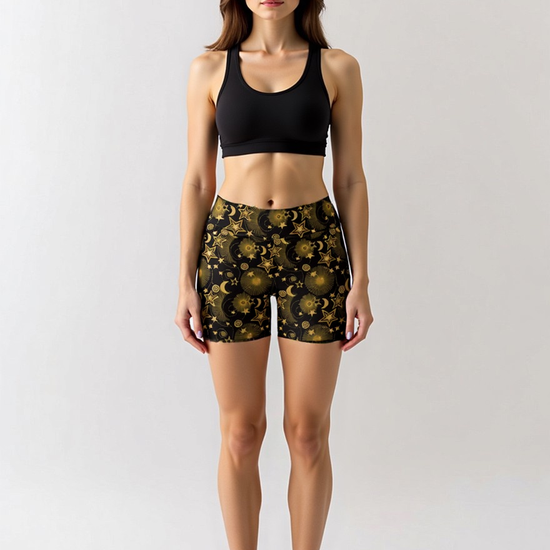 Woman wearing Midnight Halo yoga shorts with a black and gold celestial pattern of stars, suns, and moons, paired with a black sports bra, standing barefoot and turned slightly to the side against a plain light background.