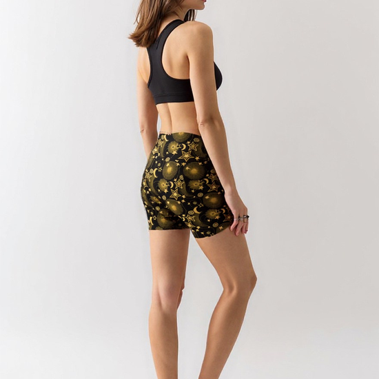 Woman wearing Midnight Halo yoga shorts with a black and gold celestial pattern of stars, suns, and moons, paired with a black sports bra, standing barefoot and turned slightly to the side against a plain light background.