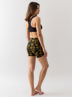 Woman wearing Midnight Halo yoga shorts with a black and gold celestial pattern of stars, suns, and moons, paired with a black sports bra, standing barefoot and turned slightly to the side against a plain light background.
