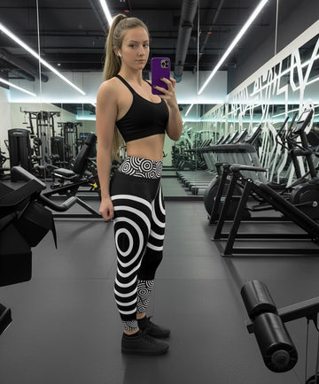 Woman taking a mirror selfie in a gym wearing black and white patterned leggings.