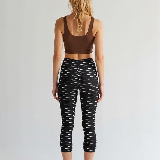 A woman is shown from the back wearing Nightfall Edit Capris, which feature a high-waisted, cropped fit with a black base and white dash print. She pairs the capri leggings with a brown sports bra, standing barefoot against a plain, light background.