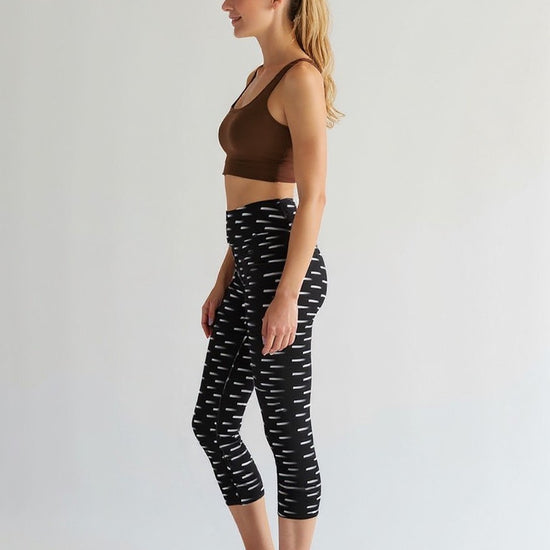 A woman is shown from the side wearing Nightfall Edit Capris, which feature a high-waisted, cropped fit with a black base and white dash print. She pairs the capri leggings with a brown sports bra, standing barefoot against a plain, light background.