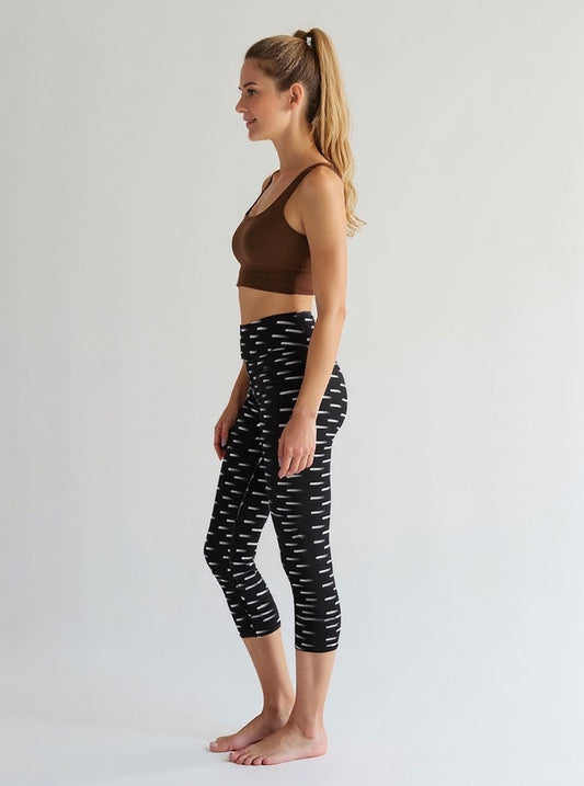 A woman is shown from the side wearing Nightfall Edit Capris, which feature a high-waisted, cropped fit with a black base and white dash print. She pairs the capri leggings with a brown sports bra, standing barefoot against a plain, light background.