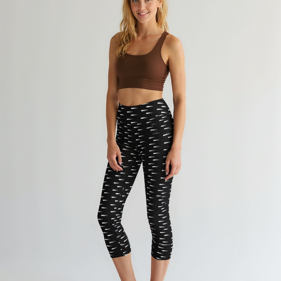 A woman stands barefoot wearing Nightfall Edit Capris, featuring a black base with an all-over white dash pattern and a high-waisted, cropped fit. She pairs the capri leggings with a brown sports bra, posing against a plain, light background.