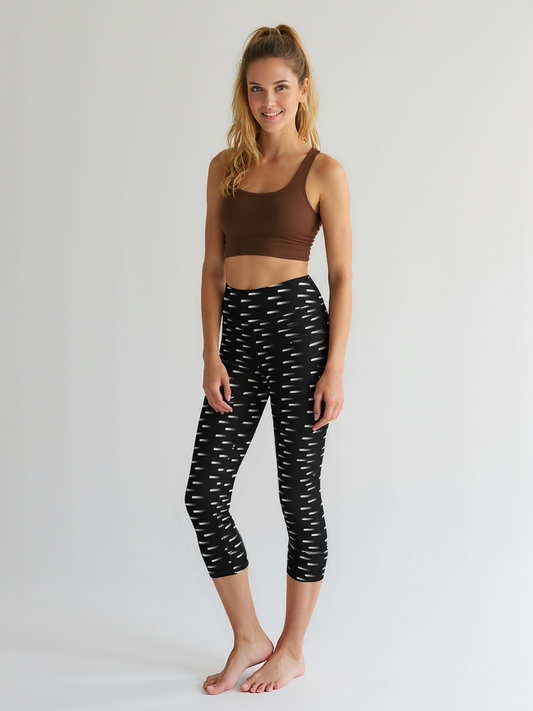 A woman stands barefoot wearing Nightfall Edit Capris, featuring a black base with an all-over white dash pattern and a high-waisted, cropped fit. She pairs the capri leggings with a brown sports bra, posing against a plain, light background.