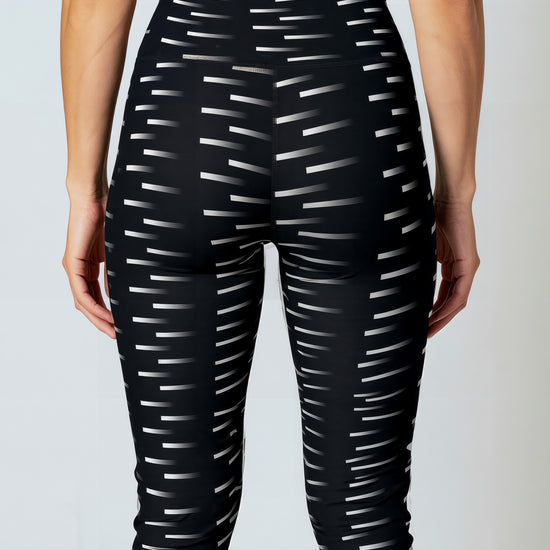 A back, close-up of a woman wearing Nightfall Edit leggings, showcasing the high-waisted fit and all-over black fabric with a white dash pattern. The image highlights the sleek waistband and the modern geometric print.
