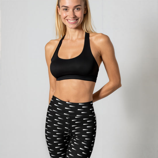 A woman stands smiling while wearing Nightfall Edit leggings, featuring a high-waisted fit and an all-over black and white dash print. She pairs the leggings with a black sports bra and black athletic shoes, posed against a plain light background.