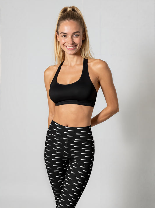 A woman stands smiling while wearing Nightfall Edit leggings, featuring a high-waisted fit and an all-over black and white dash print. She pairs the leggings with a black sports bra and black athletic shoes, posed against a plain light background.