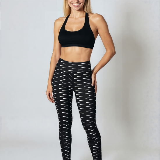 A woman stands smiling while wearing Nightfall Edit leggings, featuring a high-waisted fit and an all-over black and white dash print. She pairs the leggings with a black sports bra and black athletic shoes, posed against a plain light background.