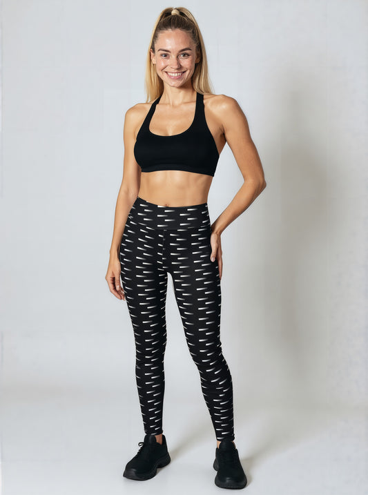 A woman stands smiling while wearing Nightfall Edit leggings, featuring a high-waisted fit and an all-over black and white dash print. She pairs the leggings with a black sports bra and black athletic shoes, posed against a plain light background.