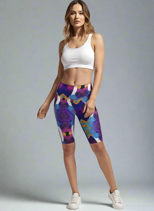 Woman wearing Ocean Mirage Biker Shorts with a vibrant abstract pattern in purple, blue, orange, and white; paired with a white sports bra and white sneakers, posing with one hand on her hip against a plain background.