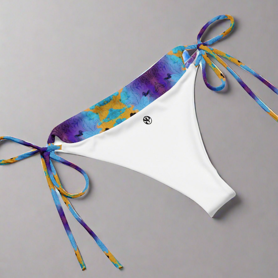 Pixie & Sherbet Ocean Mirage String Bikini bottoms laid flat on a neutral gray background. White central panel with smooth stretch fabric, vibrant tie-dye waistband in shades of blue, purple, and yellow, and matching patterned side ties in neat bows. Small black circular Pixie & Sherbet logo printed on the front, highlighting premium branding and luxe swimwear design.