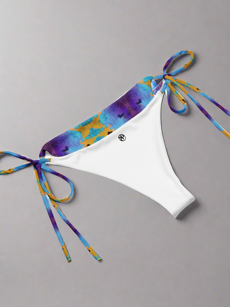 Pixie & Sherbet Ocean Mirage String Bikini bottoms laid flat on a neutral gray background. White central panel with smooth stretch fabric, vibrant tie-dye waistband in shades of blue, purple, and yellow, and matching patterned side ties in neat bows. Small black circular Pixie & Sherbet logo printed on the front, highlighting premium branding and luxe swimwear design.