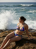 Woman in a colorful bikini sitting on a rocky beach with ocean waves in the background