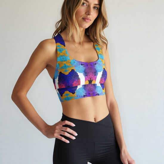 Woman wearing Ocean Mirage Longline sports bra with a vibrant abstract floral print in shades of blue, purple, yellow, and white, paired with high-waisted black leggings, standing against a plain light background.