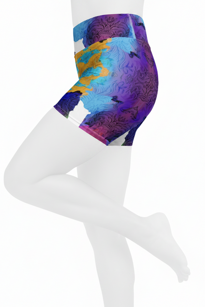 A close-up of a model's lower torso and legs wearing high-waisted athletic shorts with a colorful abstract print in purple, blue, yellow, and white on a plain white background. 