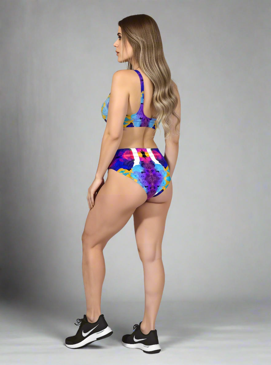 Back view of model in Pixie & Sherbet “Ocean Mirage Surf” bikini — bold multicoloured abstract print in blue, purple, yellow, and white, high-waisted bottoms and sports-bra style top, paired with black Nike sneakers, standing against a plain grey background.