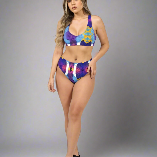 Model in Pixie & Sherbet “Ocean Mirage Surf” bikini — bold abstract print in blue, purple, red, and yellow, posing confidently in high-waisted bottoms and sports-bra style top, paired with black sneakers, against a plain grey background.