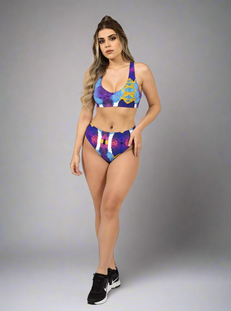 Model in Pixie & Sherbet “Ocean Mirage Surf” bikini — bold abstract print in blue, purple, red, and yellow, posing confidently in high-waisted bottoms and sports-bra style top, paired with black sneakers, against a plain grey background.