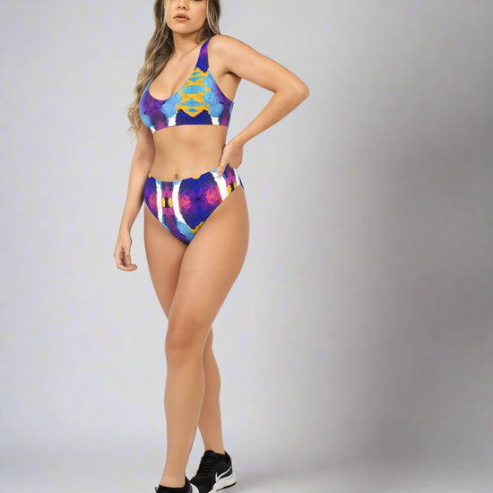 Model wearing Pixie & Sherbet “Ocean Mirage Surf” bikini — vibrant abstract print in blue, purple, yellow, and white, styled with black athletic sneakers, standing confidently against a light grey background.