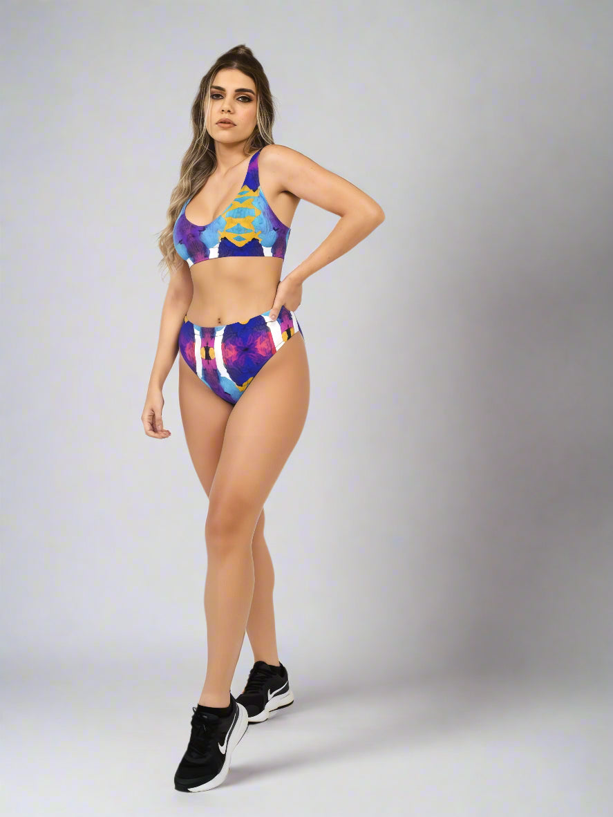 Model wearing Pixie & Sherbet “Ocean Mirage Surf” bikini — vibrant abstract print in blue, purple, yellow, and white, styled with black athletic sneakers, standing confidently against a light grey background.