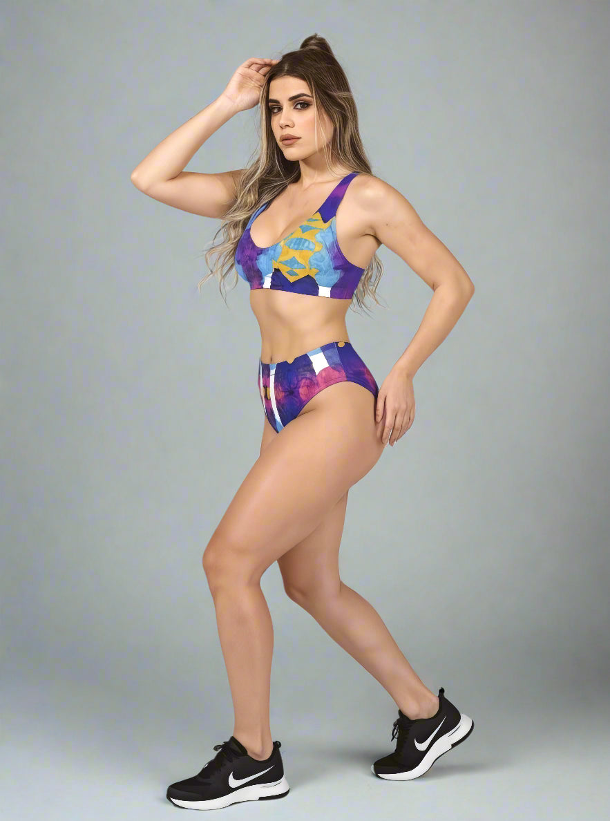 Side view of model in Pixie & Sherbet “Ocean Mirage Surf” bikini — vibrant abstract print in blue, purple, yellow, and white, high-waisted bottoms and sports-bra style top, paired with black Nike sneakers, long wavy hair, against a plain background.
