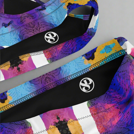 Close-up flat lay of Pixie & Sherbet “Ocean Mirage Surf” bikini — vibrant abstract print in purple, blue, yellow, black, and white, featuring sports-bra style top and matching high-waisted bottoms with detailed stitching, displayed on a plain grey background.